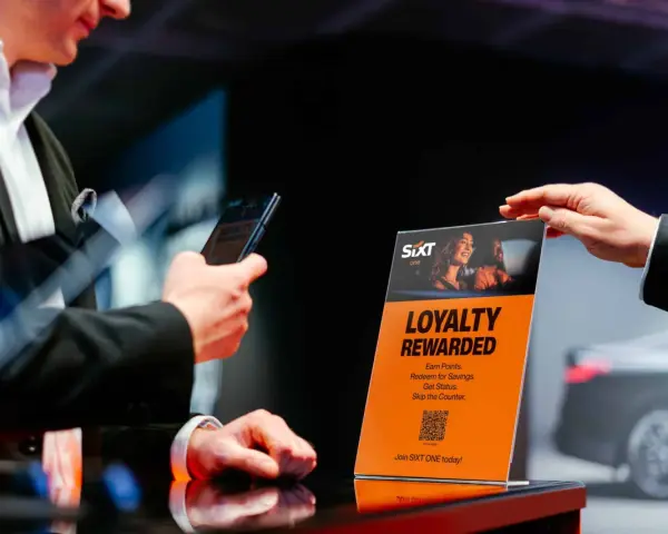 Ein Mann hält ein Smartphone, während eine Hand ein orangefarbenes Schild mit der Aufschrift "Loyalty Rewarded" und dem SIXT ONE Logo hochhält.