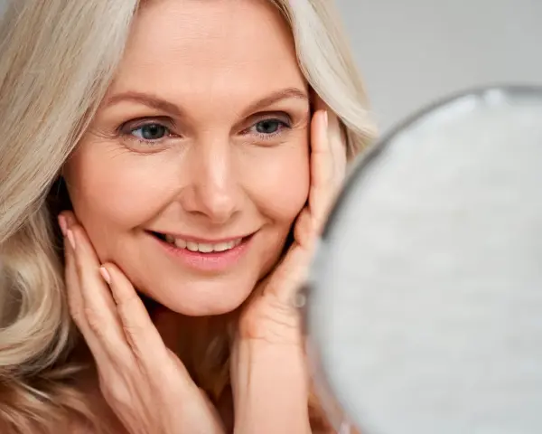 Une femme blonde sourit en se regardant dans un miroir, ses mains caressant son visage. Le maquillage paupière tombante avant après est subtil.