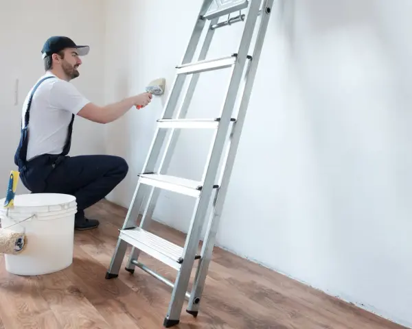 Hombre con overol azul y gorra, usando brocha para limpiar paredes. Escalera y cubo de pintura a su lado.