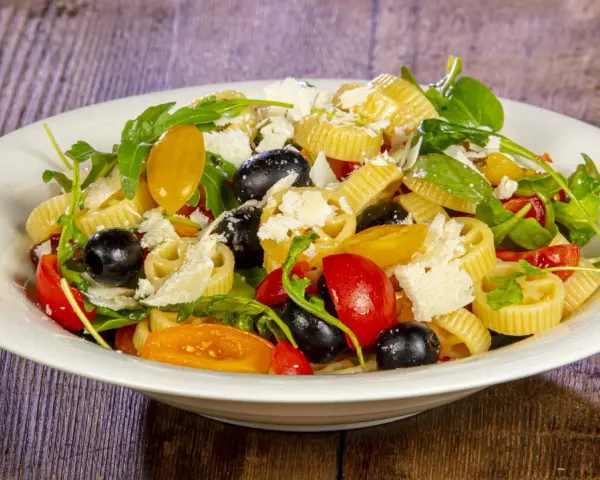 Un piatto di pasta alla crudaiola con pomodorini, olive nere, rucola e scaglie di parmigiano. Fresca e colorata.