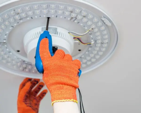 Ręka w pomarańczowej rękawicy odkręca śrubę plafonu LED. Instrukcja jak zdjąć plafon krok po kroku.