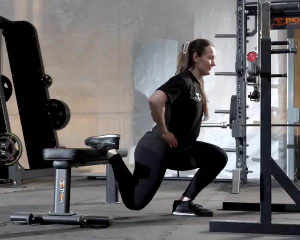 Kvinna utför en split squat i ett gym. Hennes ena fot vilar på en bänk bakom henne.