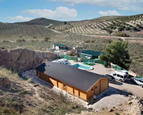 Casa de madera con piscina y caravana en terreno rústico, rodeada de olivos y colinas. Ideal para quienes buscan un refugio natural.