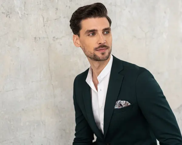 Elegancki mężczyzna w ciemnozielonej marynarce i białej koszuli z ozdobną poszetką. Jego kołnierzyk koszuli, czyli stójka, jest lekko rozpięty.