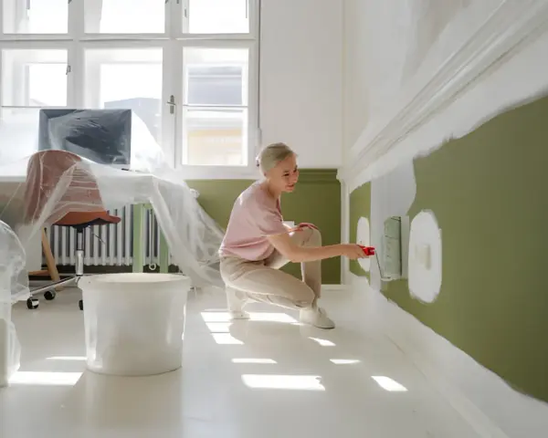 Kobieta maluje ścianę na zielono, klęcząc obok wiadra z farbą.
