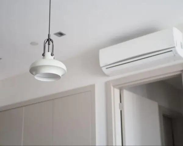 Biała klimatyzacja ścienna nad drzwiami, obok lampy wiszącej.