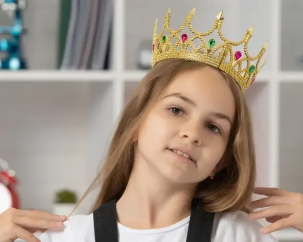 Mała egocentryczka w złotej koronie z klejnotami, pewna siebie, z uśmiechem na twarzy, na tle regału z książkami i zabawkami.
