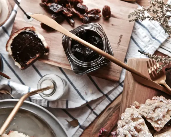 Słodkie śniadanie: kromka chleba z kremem czekoladowym, słoiczek z ciemnym dżemem, daktyle i ciastka. Idealna nutella bez orzechów!