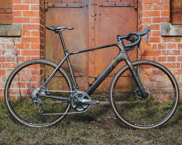 A sleek black Salsa Warroad gravel bike with orange accents stands against a rustic brick wall and weathered metal door.