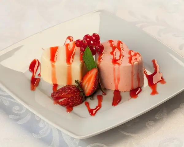 Dwie panna cotta w kształcie serc, polane sosem truskawkowym, ozdobione owocami i miętą.