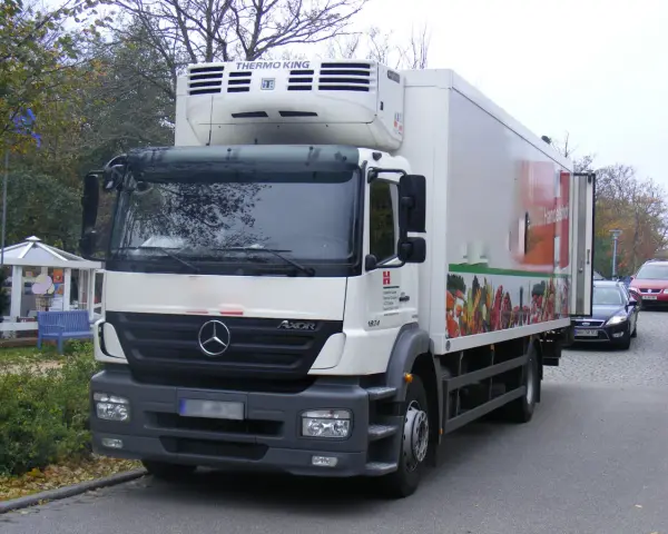 Biały Mercedes Axor z chłodnią, ozdobiony grafiką warzyw, gotowy do dostawy świeżych produktów.
