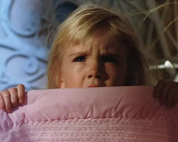 Bambina spaventata si nasconde sotto una coperta rosa, gli occhi sgranati in un'espressione di puro horror psicologico.