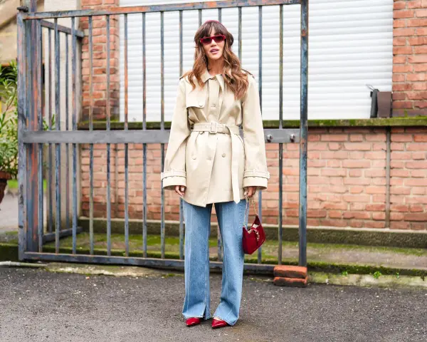 Mujer con gabardina beige y vaqueros bootcut, gafas rojas y bolso rojo.