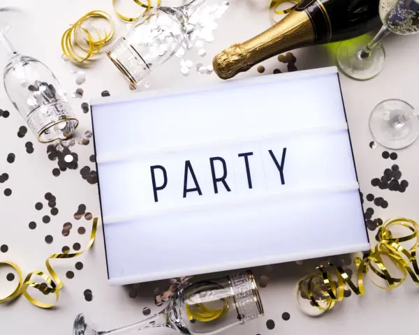 Świętowanie zaczyna się od zaproszeń! Na stole leżą kieliszki, szampan i konfetti, a tablica świetlna głosi "PARTY".