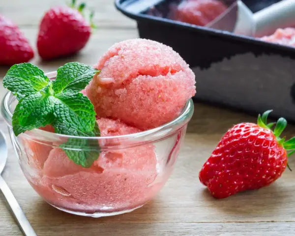 Erfrischendes Erdbeer-Sorbet-Eis in einer Glasschale, garniert mit Minze. Daneben liegen frische Erdbeeren.