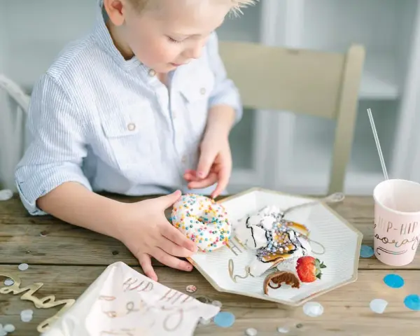 En pojke med en randig skjorta tar en donut med strössel. Det är en festlig dukning med konfetti och en mugg med texten "Hip Hip Hooray". En härlig stund av burken lek.