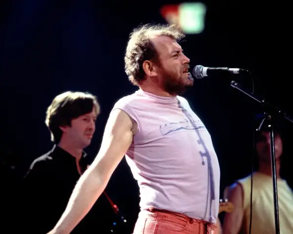 Joe Cocker śpiewa na scenie, a jego charakterystyczne utwory niosą się po sali. W tle widać innych muzyków.