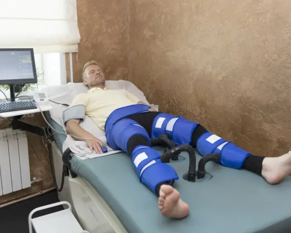 Mężczyzna leży na łóżku terapeutycznym, podłączony do aparatury do limfodrenażu. Widoczne są jego nogi w specjalnych mankietach, które mają zapewnić widoczne limfodrenaż efekty.