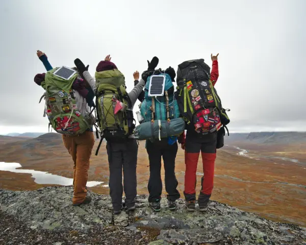 Quatre randonneurs, sacs à dos prêts, contemplent un paysage sauvage. Leur équipement, incluant des panneaux solaires, suggère une préparation pour une liste matériel randonnée 5 jours.