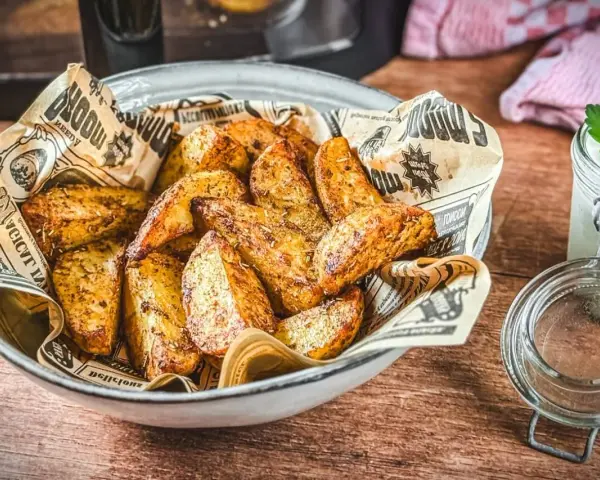 Knusprige Kartoffelecken aus dem Airfryer, serviert in einer Schüssel mit Zeitungspapier. Daneben ein Glas mit Dip und Kräutern.