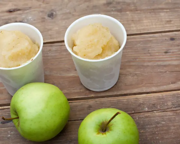 Zwei Becher mit erfrischendem Apfel Sorbet stehen neben zwei grünen Äpfeln auf einer Holzoberfläche.