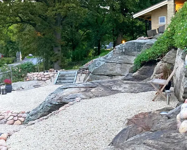 En stenlagd uteplats med grusgångar, stenmurar och trappor som leder upp mot ett hus. En inspiration för en naturlig och robust utomhusmiljö.