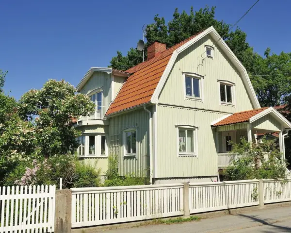 Ljusgrönt trähus med tegeltak och vit staket. En vacker trädgård med blommande buskar. Planerar att byta fasad för att fräscha upp huset.