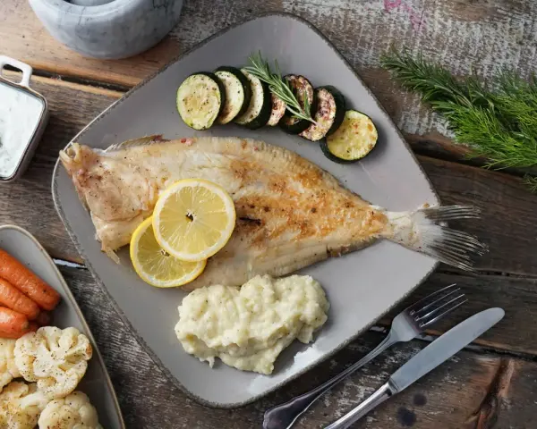 Gebratene Scholle, ein köstlicher Plattfisch, serviert mit Zucchini, Kartoffelpüree, Karotten und Blumenkohl. Zitronenscheiben und Kräuter garnieren das Gericht.