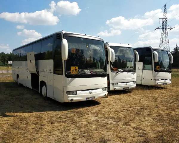 Trzy białe autobusy prywatne stoją na trawie. Jeden z nich ma otwarte drzwi. W tle las i słup energetyczny.