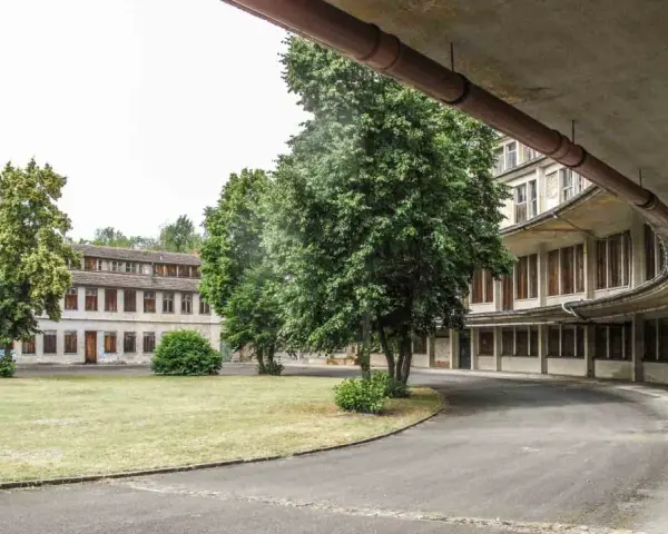 Alte Gebäude im olympisches Dorf Berlin, umgeben von Bäumen und Grünflächen. Eine Straße führt an einem geschwungenen Gebäude mit vielen Fenstern vorbei.