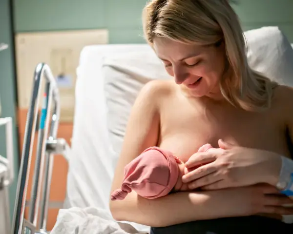 Uśmiechnięta mama karmi piersią noworodka. Pierwsze krople siary z piersi to cenny początek dla maluszka.