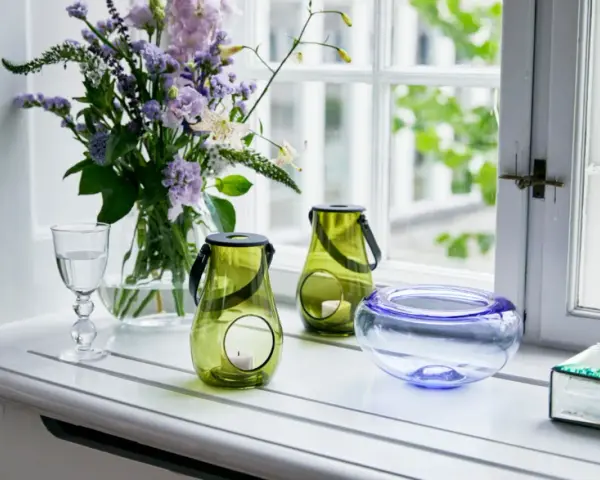 Ein Strauß lila Blumen und grüne Windlichter schmücken die Fensterbank. Ein Glas Wasser und eine blaue Schale runden die Dekoration ab.