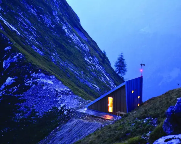 Un moderno bivacco interno, illuminato da una luce calda, si staglia su un pendio montuoso al crepuscolo.