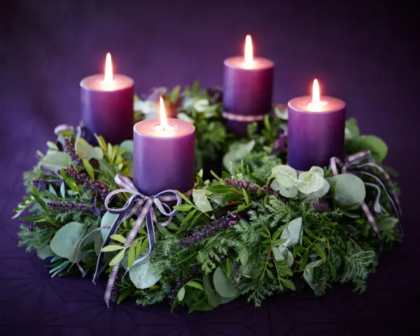 Four purple candles glow on a lush green Advent wreath, adorned with ribbons. The final candle's flame flickers, marking the 4th Advent.