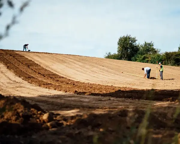 Des ouvriers travaillent sur un terrain en pente, utilisant des outils pour aplanir le sol. L'un mesure avec un mètre, l'autre pelle.