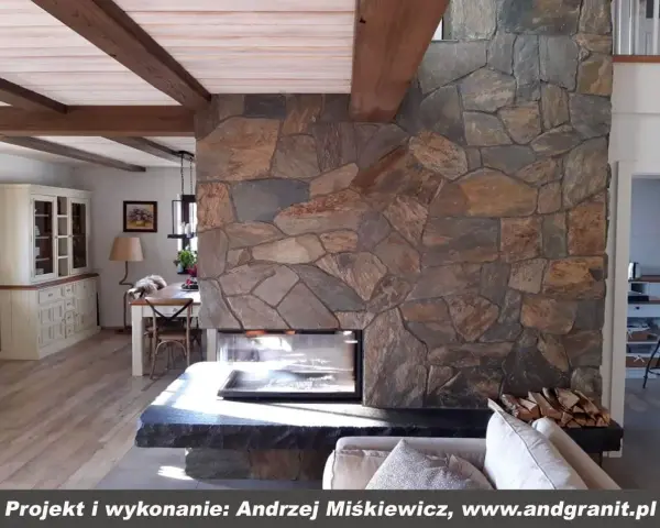 Kamienny kominek z naturalnego łupku, z drewnianymi belkami na suficie, tworzy przytulną atmosferę w salonie.