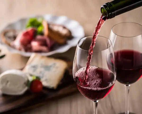 Rött vin hälls upp i glas, perfekt vin till grillat. Ost och charkuterier i bakgrunden.