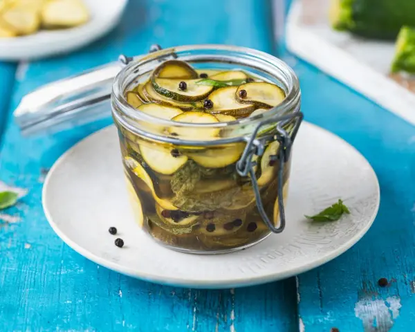 Barattolo di zucchine in agrodolce, con pepe nero e foglie di menta, su un piatto bianco appoggiato su un tavolo blu.
