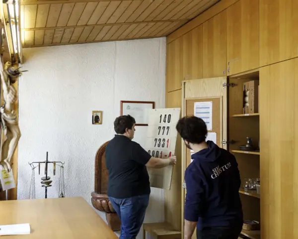Zwei Männer im katholischen Priesterseminar hängen eine Liste mit Zahlen auf. Ein Kruzifix hängt an der Wand.
