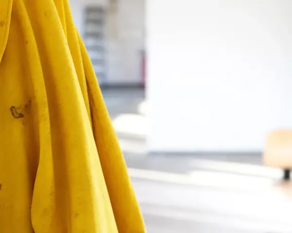 Gelbe Regenjacke hängt an einer Wand, bereit für die Regenjacke reparieren. Flecken auf dem Stoff deuten auf vergangene Abenteuer hin.