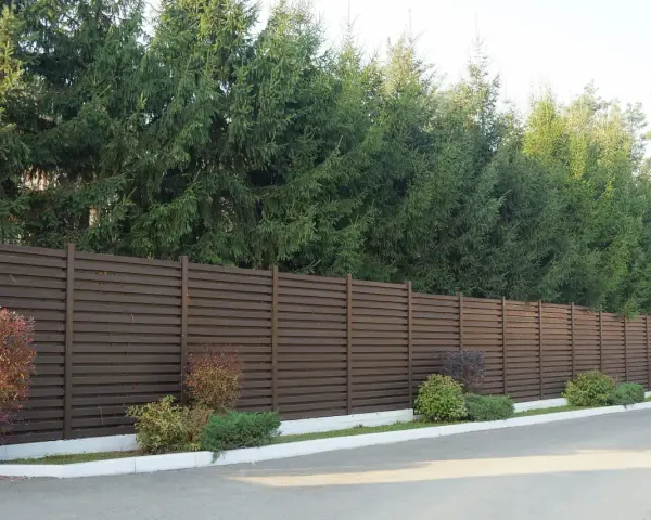 Nowoczesny, brązowy płot lamelowy to świetny sposób, by czym zasłonić płot od sąsiada. Za nim rosną wysokie świerki.