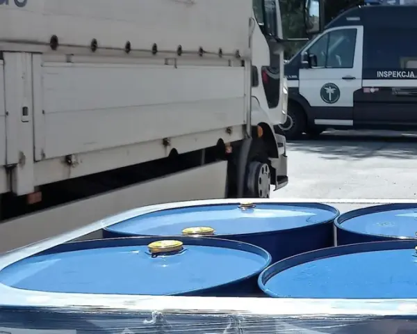 Niebieskie beczki z żółtymi zamknięciami, przygotowane do transportu towarów niebezpiecznych, obok ciężarówki i radiowozu Inspekcji Transportu Drogowego.