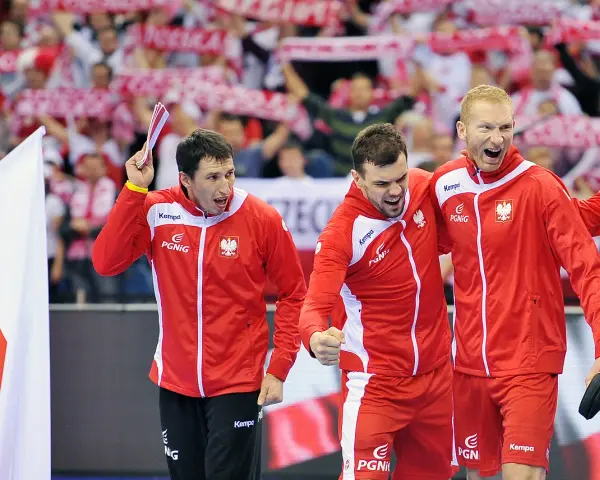 Trzech najlepszych piłkarzy ręcznych Polski w czerwonych strojach świętuje zwycięstwo. W tle kibice z biało-czerwonymi flagami.