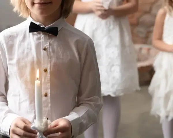 Dziecko w białej koszuli z czarną muszką trzyma zapaloną świecę. W tle inne dzieci w białych strojach.