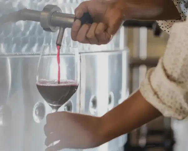 Rött vin hälls upp i ett glas från en stor tank. En hand vrider på kranen, kanske för att kontrollera tillsatser i vin.