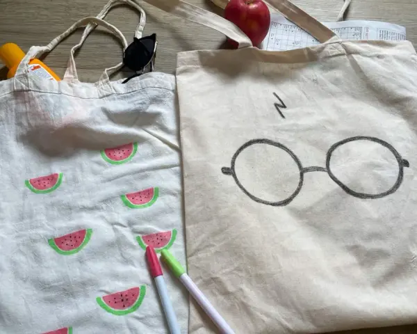 Zwei jute beutel bemalen: einer mit Wassermelonen, der andere mit Harry Potters Brille. Sonnencreme, Sonnenbrille und Stifte liegen daneben.