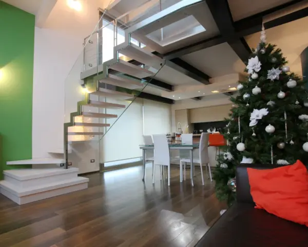 Un moderno soppalco casa con scala in vetro e legno, albero di Natale addobbato e un divano con cuscino arancione.
