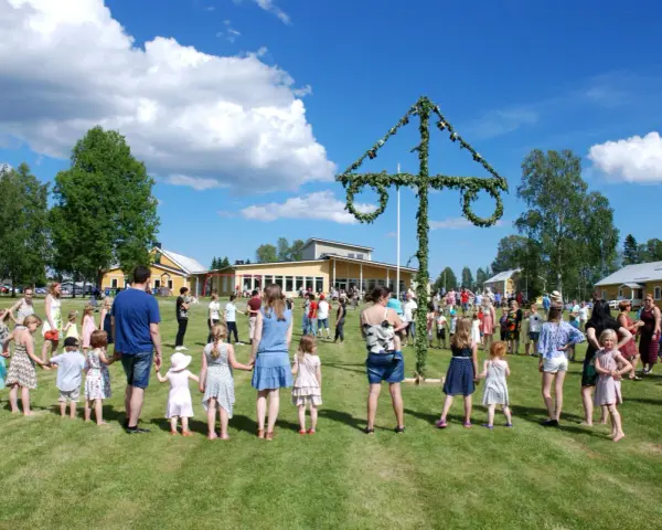 Många människor, både vuxna och barn, håller varandra i händerna och dansar runt en midsommarstång. De firar midsommar i en grönskande park med gula byggnader i bakgrunden.