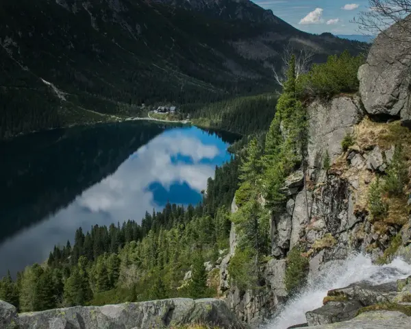 Widok z góry na Morskie Oko, odbijające chmury. Trasa turystyczna prowadzi przez skaliste zbocza z wodospadem.