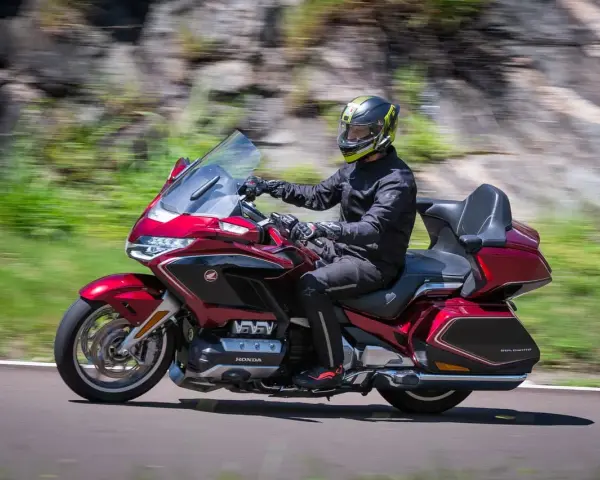 Mężczyzna w kasku jedzie lśniącym, czerwonym motocyklem uniwersalnym. W tle rozmyte zielone wzgórza i skały.